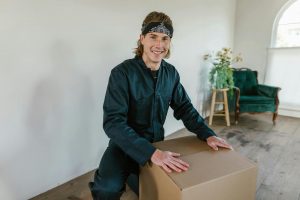 Hombre sonriente con ropa de trabajo sujetando una caja de cartón en el interior de un local.