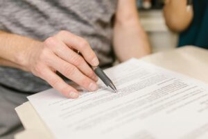 Alt: Mano sosteniendo un bolígrafo señalando un documento impreso sobre una mesa durante la revisión de un contrato o informe.