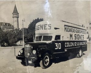Camión antiguo de mudanzas y guardamuebles “M. Signes” estacionado en una calle de Valencia, con un edificio histórico y torre al fondo en una fotografía en blanco y negro.