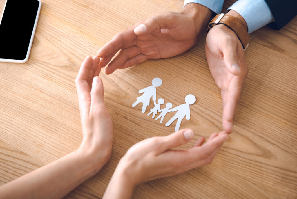 manos de dos personas protegiendo figuras de papel que representan una familia sobre una mesa