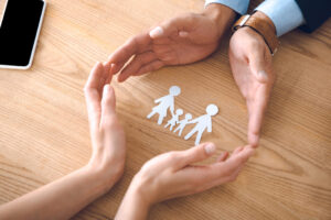 manos de dos personas protegiendo figuras de papel que representan una familia sobre una mesa
