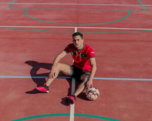 Jugador de fútbol sentado en una pista deportiva con camiseta roja y balón, en una cancha al aire libre.