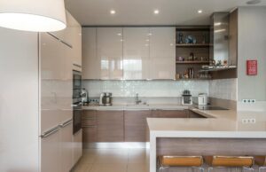 Cocina moderna de diseño minimalista con muebles en tonos beige y madera, encimera amplia con barra, electrodomésticos integrados y estanterías decorativas abiertas.