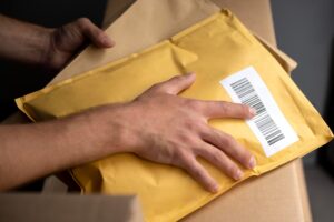 Primer plano de una persona sosteniendo un sobre acolchado amarillo con etiqueta de código de barras sobre una caja de cartón.