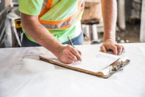 Trabajador con chaleco de seguridad tomando notas en un plano técnico sobre una mesa de trabajo en obra o taller.