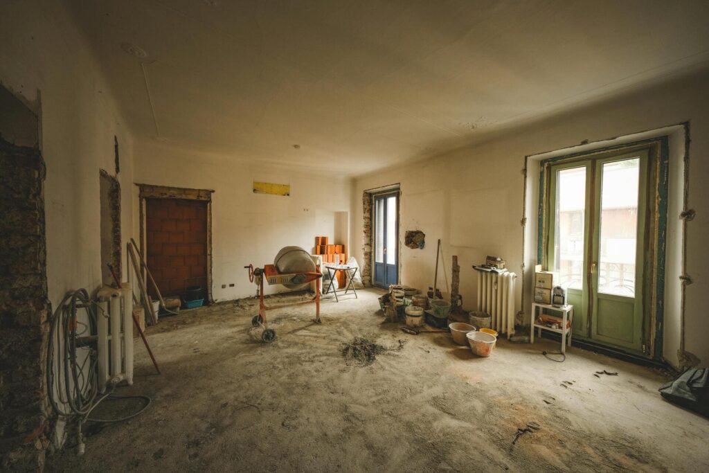 Estancia interior en proceso de reforma con suelo sin terminar, herramientas de obra, hormigonera y materiales de construcción distribuidos por la habitación.