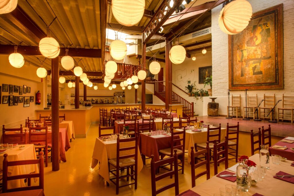 Interior de restaurante amplio y acogedor con mesas vestidas, sillas de madera, iluminación cálida con lámparas colgantes y zona de escenario al fondo.