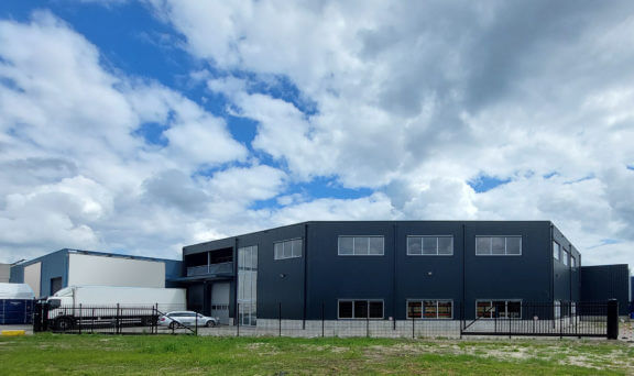 Nave industrial moderna de estructura metálica negra con grandes ventanales, zona de carga para camiones y vallado exterior bajo un cielo parcialmente nublado.