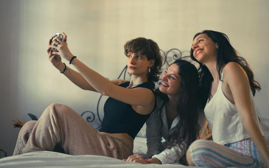 Tres jóvenes sentadas en una cama haciéndose un selfie con una cámara y sonriendo en un ambiente informal.