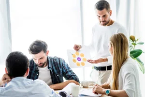 Personas reunidas en una oficina durante una reunión de trabajo, mientras un profesional presenta una estrategia o proyecto mostrando un gráfico al resto del equipo.