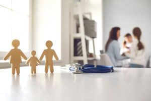 Figuras de madera de una familia (madre, hijo y padre) junto a un fonendoscopio sobre una mesa blanca, con una familia desenfocada al fondo en una consulta médica.
