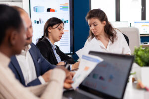 Reunión de trabajo en una oficina con pantalla de gráficos al fondo: dos mujeres revisan un informe impreso con datos y un hombre consulta un portátil abierto sobre la mesa