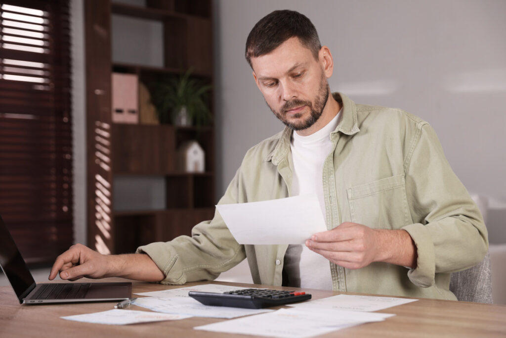 Hombre revisando documentos financieros en un escritorio con un portátil y una calculadora