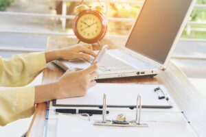 Persona utilizando un móvil junto a un portátil, un despertador y documentos sobre la mesa.