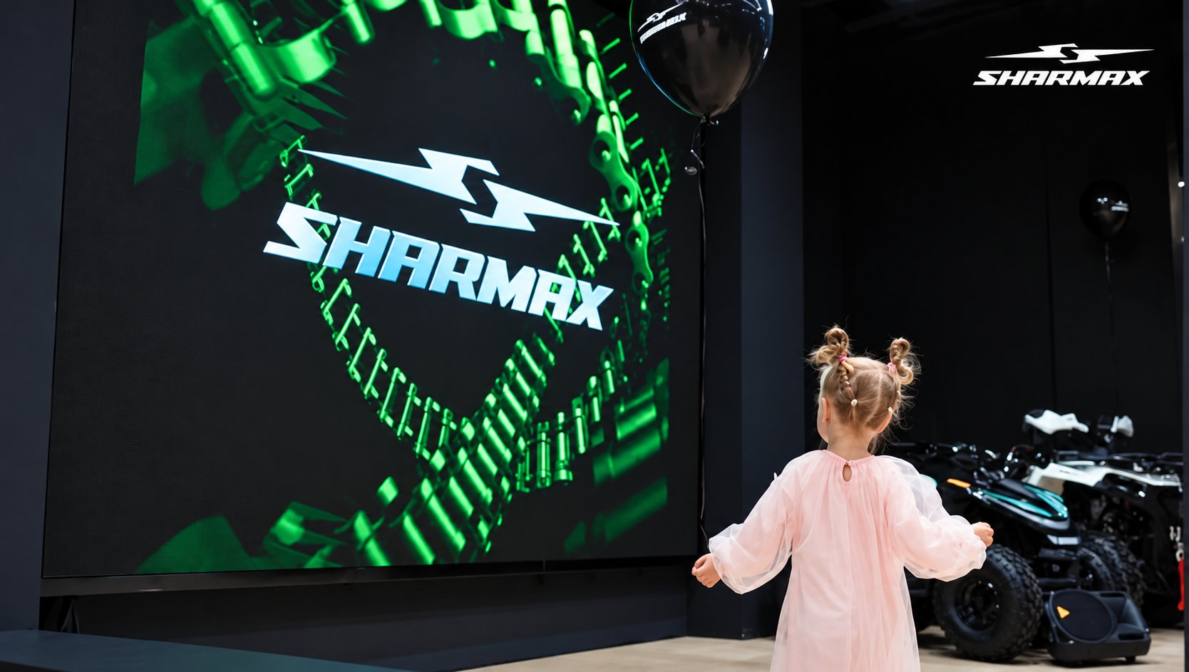 Una niña frente a una pantalla con el logotipo de Sharmax y vehículos expuestos al fondo.